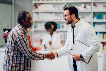 Male pharmacist shaking hands with a senior african american man patient in pharmacy