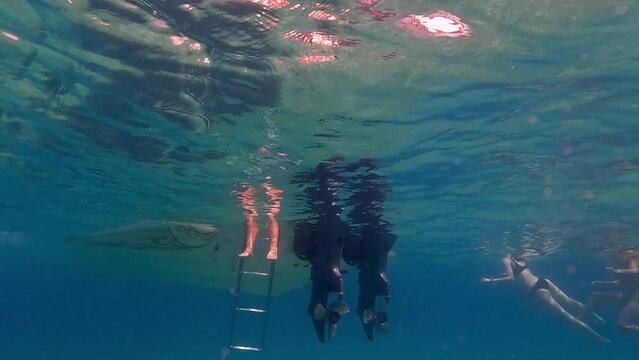 Slow motion under water scene of human legs and feet in sea water beneath surface protruding from motorboat ladder