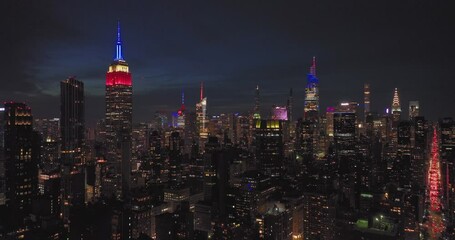 Aerial flying towards illuminated NYC skyscrapers at night with colorful famous New York City building lit red white blue 4th of July - Powered by Adobe