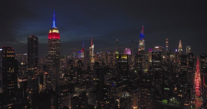 Aerial flying towards illuminated NYC skyscrapers at night with colorful famous New York City building lit red white blue 4th of July