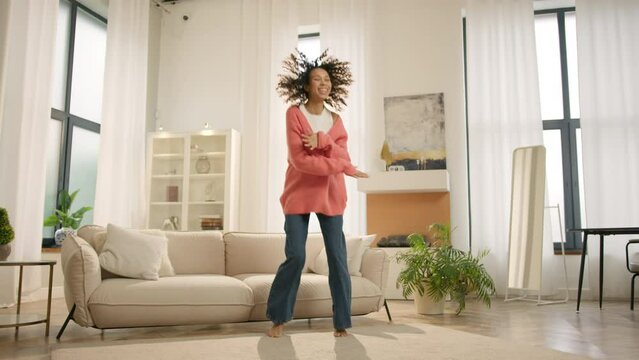 Smiling woman of color dancing for camera indoors. Cheerful mixed race female person moving hands to beat of music at home party 4K. Slow motion cute girl with curly afro hair having fun alone at home