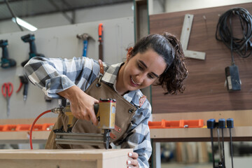 Young woman carpenter using pneumatic nail gun making new furniture at wood workshop. Young joiner...