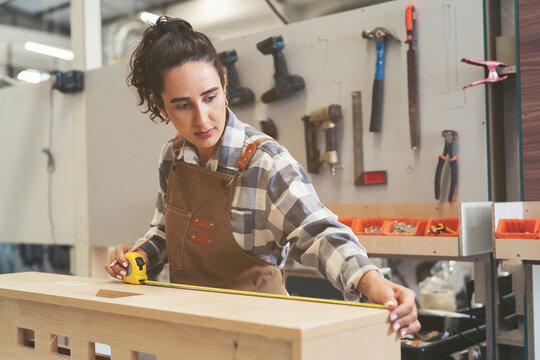 Young woman carpenter using measuring tape at carpentry workshop. Joiner female working with tape measure in wood factory