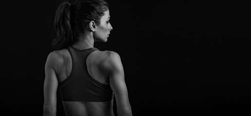 Sporty slim woman with muscular back posing in sport bra, standing on dark black shadow black background with empty copy space. Back view with profile face. Closeup banner.