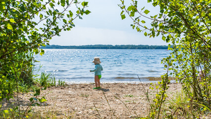 A toddler stands among green bushes on the shore of Cospuden Lake near Leipzig in summer
