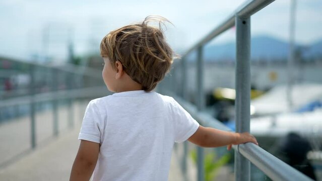 Back of child walking in bridge at bay outside during sunny beautiful summer day. One little boy enjoying vacations holding on metal handrail