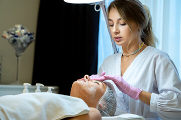 Woman cosmetologist apply white mask on woman's face, aesthetic clinic, skin care.