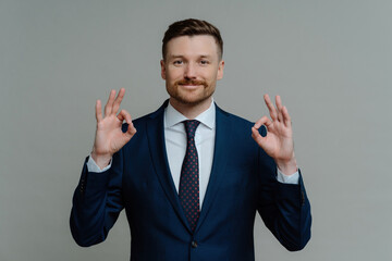 Successful businessman in formal wear, showing ok sign, smiling at camera against grey background, satisfied with work