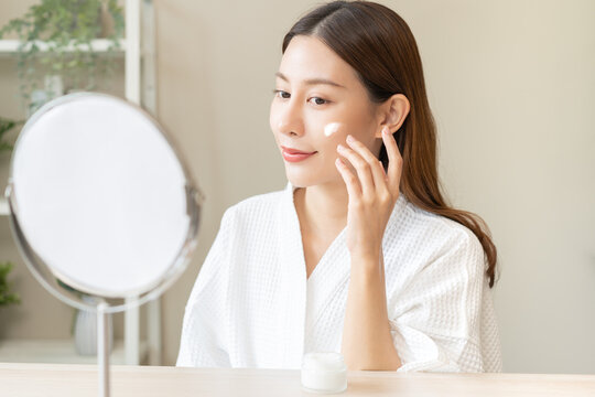 Facial beauty skin care, smile of pretty asian young woman in bathrobe looking at mirror, hand applying moisturizer lotion on her face, putting cream treatment before makeup cosmetic routine at home.