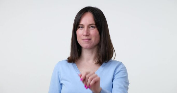 Caucasian woman stands on a white background and shakes her index finger, politely refusing someone or something. She wears a slight smile on her face, indicating a friendly demeanor.