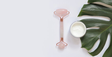 Rose quartz face massage roller and face cream. Massage roller on monstera leaf, white background, top view. The concept of natural treatment. Flat lay. Facial beauty massage, spa self-care concept © Mariia