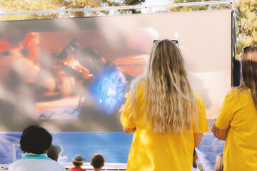 A fair-haired girl in a yellow T-shirt is watching a movie in a summer cinema.