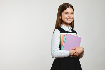 Excited girl in school uniform holding colorful books while smiling and looking at camera, standing...