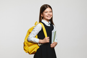 Carefree pupil girl with yellow backpack smiling and embracing notebooks during studies against white background. Back to school concept