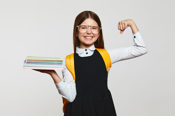 Preteen girl in school uniform and yellow backpack, holding books and showing hand biceps muscles,...