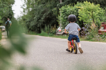 A cheerful little boy rides a running bike in a helmet outdoors. A happy child is engaged in an...