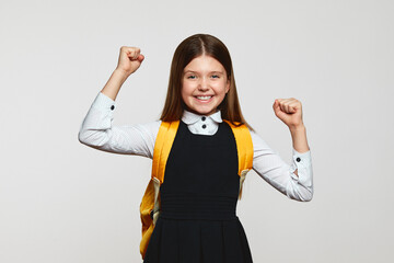 Happy little girl in school uniform and yellow backpack, celebrating success and victory standing with raised hands isolated on white background