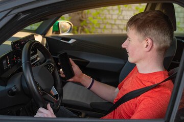 A Young Caucasian male driver sitting in the car, wearing orange t-shirt and holding smartphone in his hand. A man is driving an electric car in the summertime using car sharing.