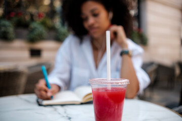 Close-up of juice cup with blurred woman writing on notebook.