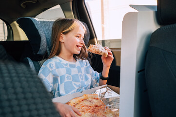 Portrait of little 5 YO girl eating just cooked Italian pizza sitting in child car seat on car back seat and looking out window . Happy childhood, fast food eating, or auto journey lunch break image