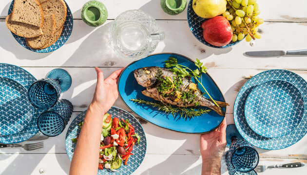 Gilt-head bream also known as Orata grilled on a barbeque grill served by a woman on a white wooden table with vegetable salad and fruits. Healthy seafood preparation concept photo.