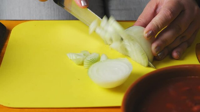Women's hands cut onion for cholent with a large knife on a plastic board.