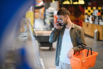 Honey, what do you want to eat. Portrait of a handsome male customer talking on his phone while shopping at the groceries standing near his shopping cart full of delicious food