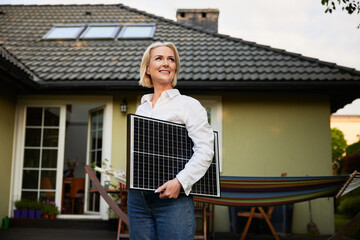 Smiling adult woman standing with solar panel outdoors at backyard of her house
