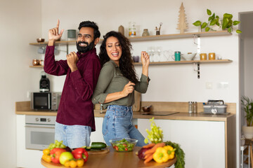 Joyful arab man and woman in casual outfits having fun while cooking together at home, dancing and singing in kitchen