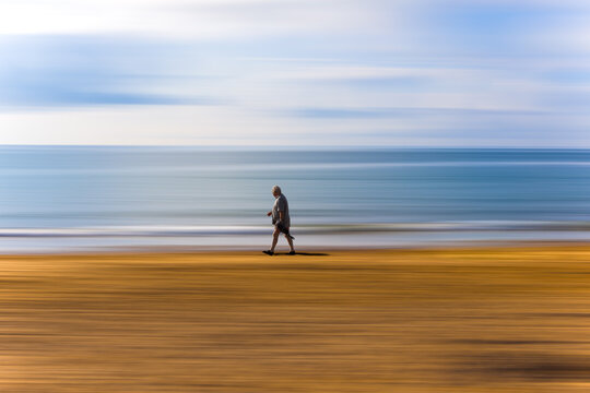 Lonely walk.  A man walking alone along the sea shore on Freshwater East sandy beach, Wales.  The background has a path blur.  Blue sea and cloudy sky.