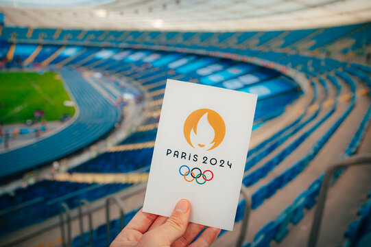 PARIS, FRANCE, JULY 7, 2023: Embracing the Symbol: Athlete Holds Iconic Emblem of Paris 2024 Summer Olympics, with Modern Stadium as a Majestic Backdrop. A Symbolic Prelude to Paris 2024 Olympics