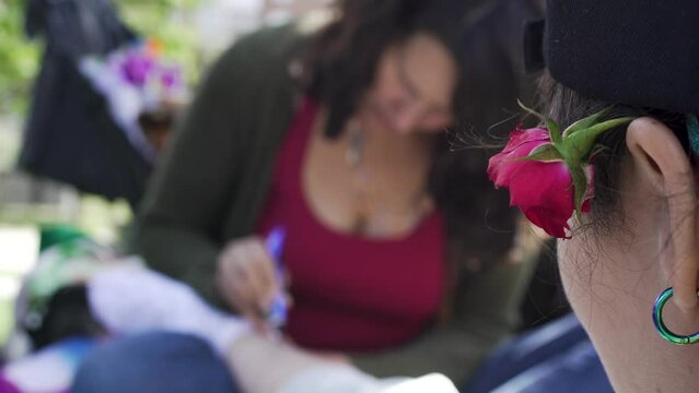 Close up of red rose in ear in foreground with background of unrecognisable woman painting a friend's leg in henna in a picnic party