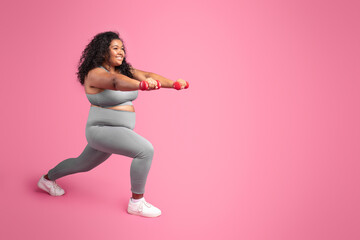 Black chubby woman holding dumbbells and making lunges exercise, training legs over pink studio...