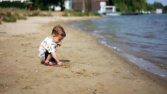 Lovely dark-haired toddler sits on the sandy shore. Kid touches wet sand and looks at water.