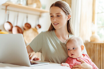 Young smiling woman, freelancer holding cute baby using laptop working online from home, watching...