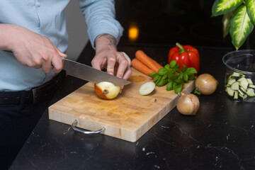 Man in blue shirt cutting and slicing onion og wooden cutting board, with other vegetables in the background.