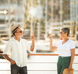 Senior Spouses Pointing Fingers Up Posing At Pier In Marina