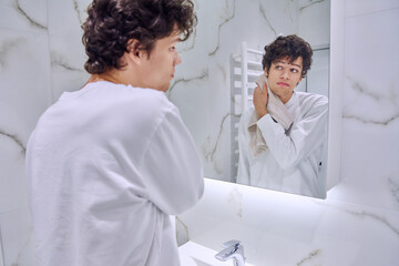 Fototapeta premium Young guy in bathroom with towel looking at his reflection in mirror