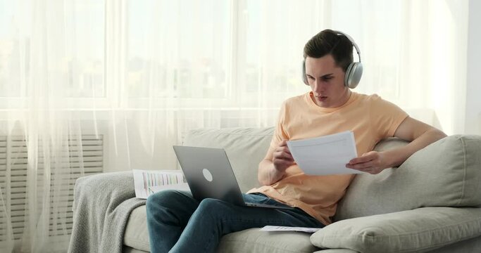 Focused man, donning a pair of headphones, immerses in work on the comfort of sofa. With dedicated gaze and a laptop positioned strategically, he delves into his tasks with unwavering determination.