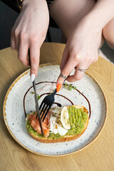 Toast with salmon and a poached egg on an avocado spread. Female hands with a fork