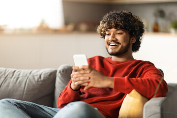 Cheerful Indian Man Messaging On Smartphone While Relaxing On Couch At Home