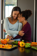 Happy biracial lesbian couple hugging and chopping vegetables in kitchen