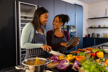 Happy biracial lesbian couple chopping vegetables using tablet in kitchen