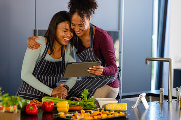 Happy biracial lesbian couple preparing meal using tablet in kitchen