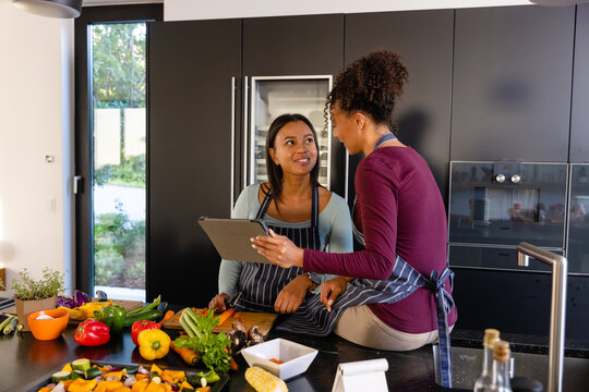 Happy biracial lesbian couple preparing meal using tablet in kitchen