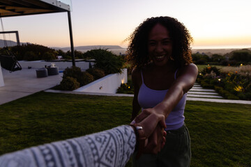 Happy biracial lesbian couple holding hands in garden at sunset