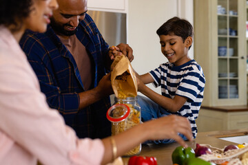 Happy biracial parents and son unpacking groceries and pouring pasta into storage jar in kitchen