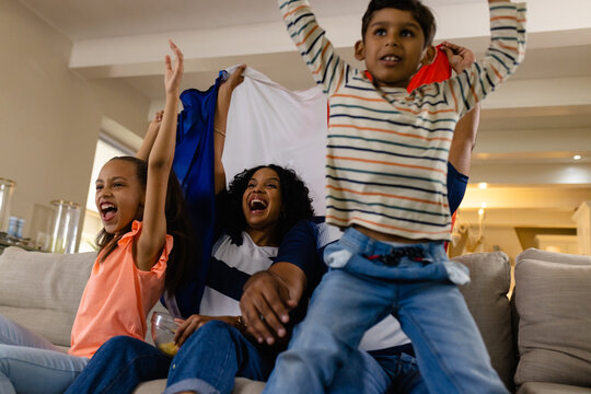 Happy biracial family sitting on couch holding french flag, watching tv and cheering - Powered by Adobe