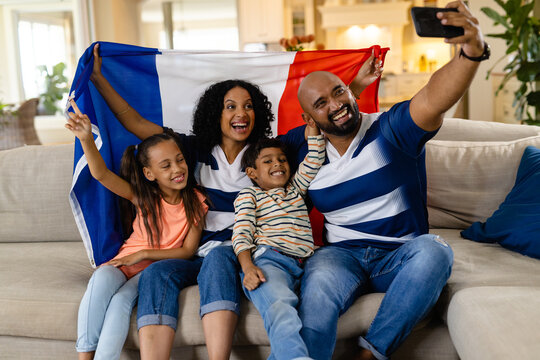 Happy biracial family sitting on couch holding french flag, taking selfie and smiling