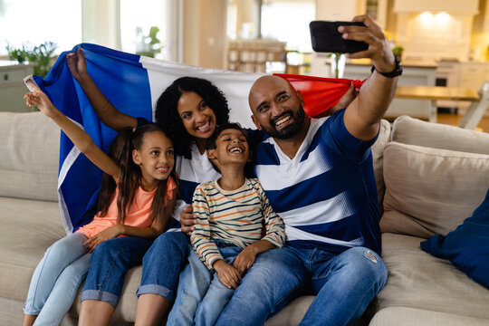 Happy biracial family sitting on couch holding french flag, taking selfie and smiling